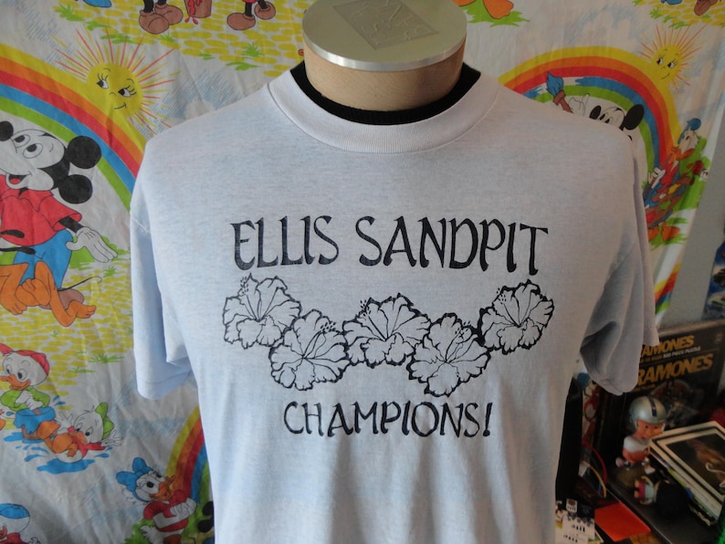 May include: Light blue t-shirt with the text "ELLIS SANDPIT CHAMPIONS!" in black, along with a floral design. The shirt has a crew neck and short sleeves. The shirt is displayed on a mannequin.