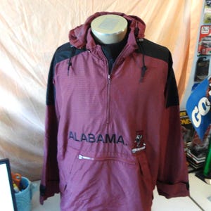 May include: A maroon and black pullover windbreaker jacket featuring a hood with drawstrings, a half-zip front closure, and a large front pocket with a zipper. The word ALABAMA is printed in black across the chest with a small mascot graphic.
