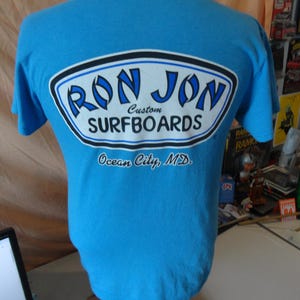 May include: A light blue t-shirt featuring the text "RON JON SURFBOARDS" in a white, black, and blue oval design. The text "Ocean City, MD" is printed below the logo. The shirt is displayed on a mannequin.
