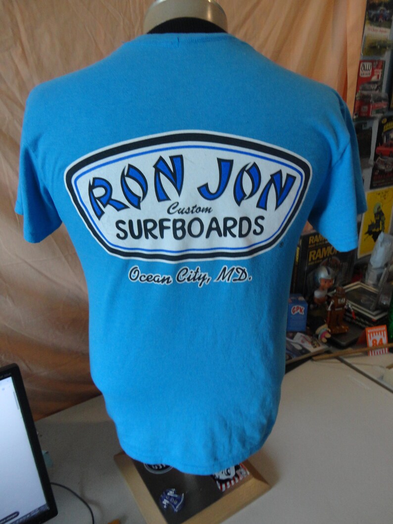 May include: A light blue t-shirt featuring the text "RON JON SURFBOARDS" in a white, black, and blue oval design. The text "Ocean City, MD" is printed below the logo. The shirt is displayed on a mannequin.