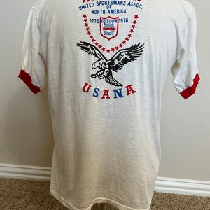 K&ouml;nnte beinhalten: Wei&szlig;es T-Shirt mit rotem Ringel. Die R&uuml;ckseite des Shirts zeigt eine Grafik eines Adlers mit dem Text "United Sportsmans Assoc. of North America 1776 Freedom 1976" und "USANA" unterhalb des Adlers.