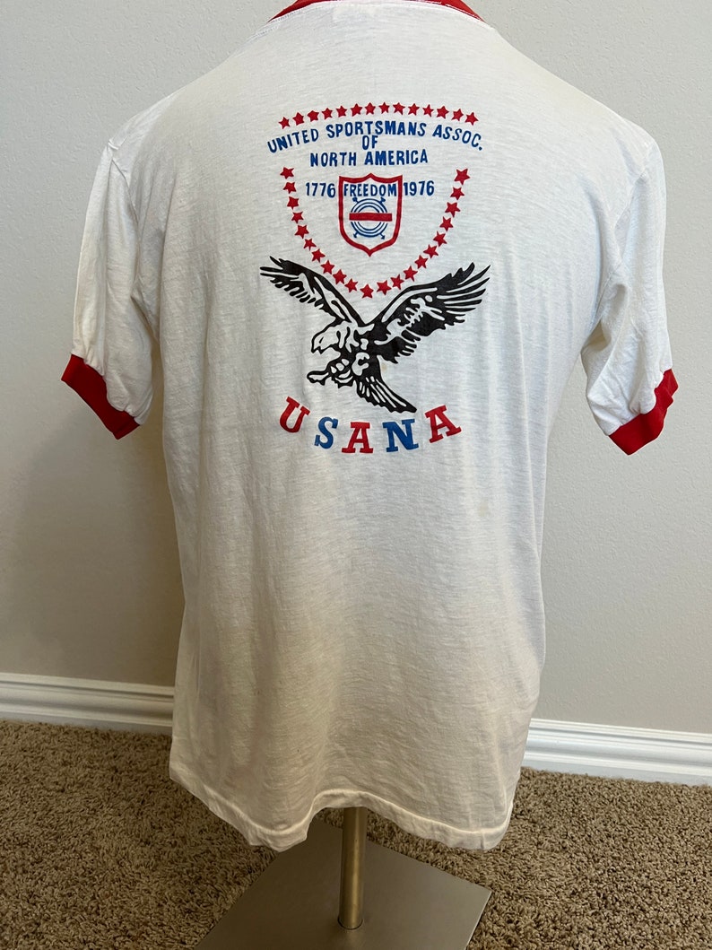 K&ouml;nnte beinhalten: Wei&szlig;es T-Shirt mit rotem Ringel. Die R&uuml;ckseite des Shirts zeigt eine Grafik eines Adlers mit dem Text "United Sportsmans Assoc. of North America 1776 Freedom 1976" und "USANA" unterhalb des Adlers.