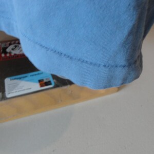 May include: A light blue t-shirt, with the bottom hem visible, rests on a wooden frame. The frame holds a black object with a label that has text and a blue and white design.
