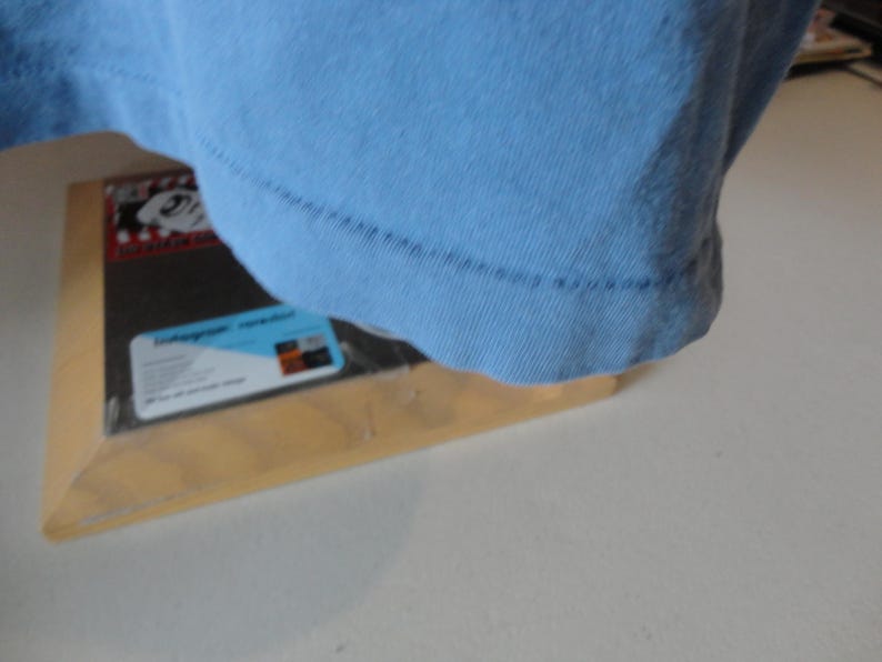 May include: A light blue t-shirt, with the bottom hem visible, rests on a wooden frame. The frame holds a black object with a label that has text and a blue and white design.