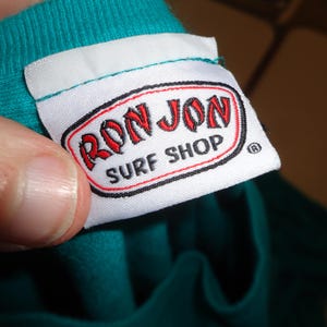 May include: A white label with the text "RON JON SURF SHOP" in red and black. The label is attached to a teal garment. The label is being held by a person.