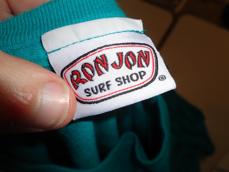 May include: A white label with the text "RON JON SURF SHOP" in red and black. The label is attached to a teal garment. The label is being held by a person.