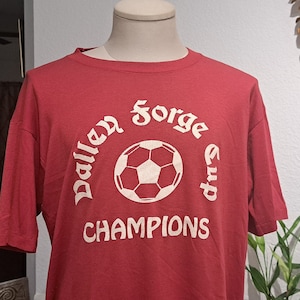 May include: Red t-shirt with a white graphic of a soccer ball and the text "Valley Forge Cup Champions".