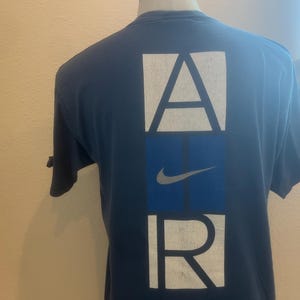 Puede incluir: Una camiseta azul marino con un gr&aacute;fico blanco de "AIR" en la espalda. La "A" y la "R" est&aacute;n en cajas, y la "I" es una caja azul con un swoosh blanco de Nike.