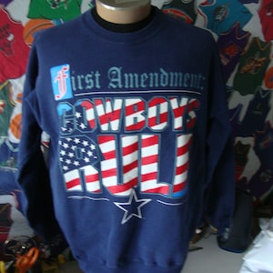 Puede incluir: Una sudadera azul marino con un diseño de bandera americana roja, blanca y azul. El texto "First Amendment: Cowboys Rule" está impreso en la parte delantera de la sudadera.