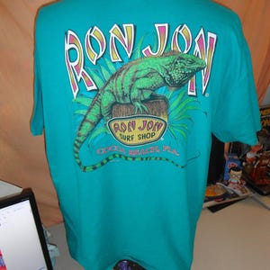 May include: Teal t-shirt with a graphic of a green iguana on a wooden post, with the text "RON JON" above and "COCOA BEACH, FLA." below. The shirt is displayed on a mannequin.