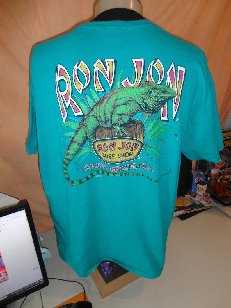 May include: Teal t-shirt with a graphic of a green iguana on a wooden post, with the text "RON JON" above and "COCOA BEACH, FLA." below. The shirt is displayed on a mannequin.