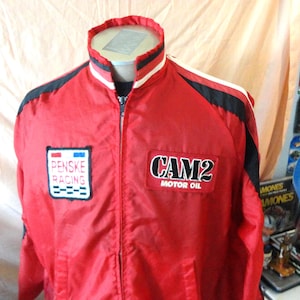 May include: A red zip-up racing jacket featuring black and white accents on the collar and shoulders. The chest displays two embroidered patches: a white Penske Racing logo on the right and a black and white CAM2 Motor Oil logo on the left.