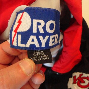 Pode incluir: Um close-up de uma etiqueta azul Pro Player com texto branco e um design de raio vermelho. A etiqueta est&aacute; presa a uma pe&ccedil;a de roupa vermelha e preta. A etiqueta diz "GD/L (2) PIECES MADE IN KOREA ECHO EN KOREA".