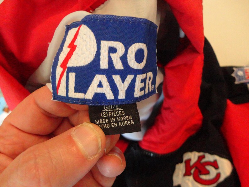 Pode incluir: Um close-up de uma etiqueta azul Pro Player com texto branco e um design de raio vermelho. A etiqueta est&aacute; presa a uma pe&ccedil;a de roupa vermelha e preta. A etiqueta diz "GD/L (2) PIECES MADE IN KOREA ECHO EN KOREA".