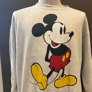 May include: A light gray sweatshirt featuring a large graphic of Mickey Mouse. Mickey is depicted in his classic pose, with a black head and ears, a white face, red shorts, yellow shoes, and black gloves. The sweatshirt has a crew neck and long sleeves.