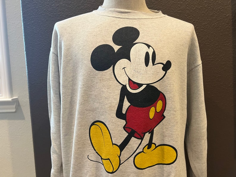 May include: A light gray sweatshirt featuring a large graphic of Mickey Mouse. Mickey is depicted in his classic pose, with a black head and ears, a white face, red shorts, yellow shoes, and black gloves. The sweatshirt has a crew neck and long sleeves.