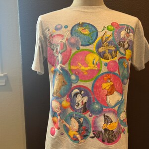 Puede incluir: Una camiseta gris claro con un estampado colorido de personajes de Looney Tunes. El diseño incluye Bugs Bunny, Piolín, el Pato Lucas y otros, dentro de marcos circulares vibrantes. La camiseta tiene mangas cortas.