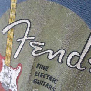May include: Close-up of a blue t-shirt featuring the Fender logo in white, with a red and white electric guitar graphic. The text "FINE ELECTRIC GUITARS" is also visible.
