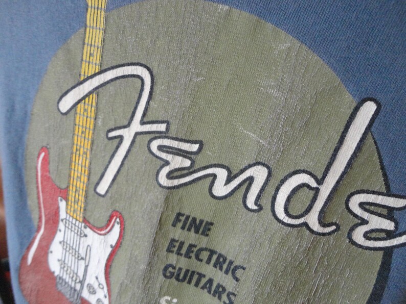 May include: Close-up of a blue t-shirt featuring the Fender logo in white, with a red and white electric guitar graphic. The text "FINE ELECTRIC GUITARS" is also visible.
