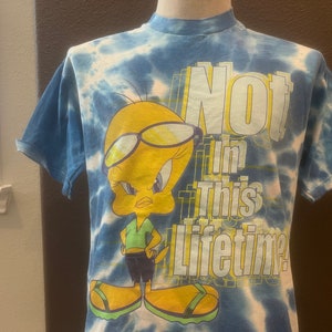May include: A blue and white tie-dye t-shirt featuring Tweety Bird wearing glasses and the phrase "Not In This Lifetime." The cartoon character is in a green shirt, black shorts, and yellow sandals. The shirt has short sleeves.