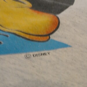 May include: Close-up of a white t-shirt with a blue and yellow cartoon character design. The shirt has a blue and yellow design with a black outline. The text "&copy; DISNEY" is printed on the bottom right corner of the shirt.