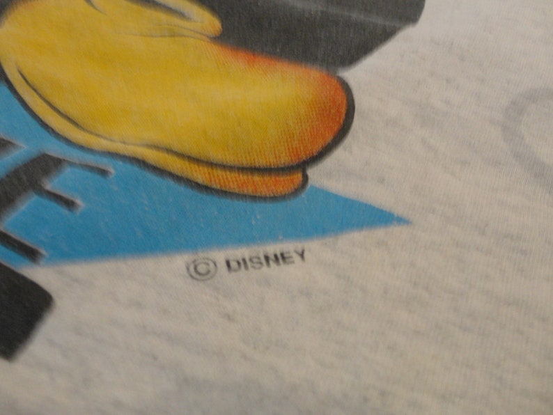 May include: Close-up of a white t-shirt with a blue and yellow cartoon character design. The shirt has a blue and yellow design with a black outline. The text "&copy; DISNEY" is printed on the bottom right corner of the shirt.