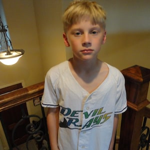 May include: A white baseball jersey with the text "DEVIL RAYS" in green and blue. The jersey has a button-up front and short sleeves with navy blue trim. The person is standing on a staircase.