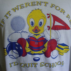 May include: White t-shirt with a cartoon duck wearing a red and blue striped shirt. The duck is surrounded by various sports equipment, including a tennis racket, a basketball, a bowling ball, and a hockey puck. The text "If it weren't for P.E... I'd quit school" is printed on the shirt.
