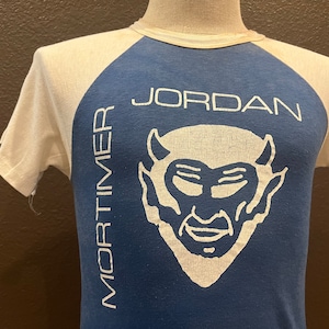 May include: A white and blue baseball tee shirt with a white devil graphic and the text "MORTIMER JORDAN" printed on the front.