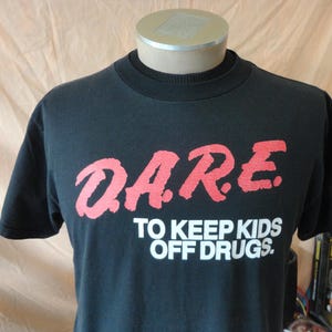 May include: Black t-shirt with the D.A.R.E. logo in red and the text "TO KEEP KIDS OFF DRUGS" in white. The shirt is displayed on a mannequin.