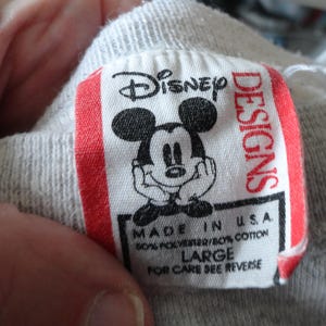 May include: A close-up of a gray garment tag with the Disney logo and a smiling Mickey Mouse illustration. The tag reads "Disney Designs" and "Made in U.S.A." with fabric content and size "LARGE".