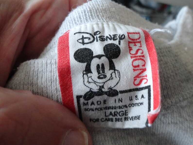 May include: A close-up of a gray garment tag with the Disney logo and a smiling Mickey Mouse illustration. The tag reads "Disney Designs" and "Made in U.S.A." with fabric content and size "LARGE".