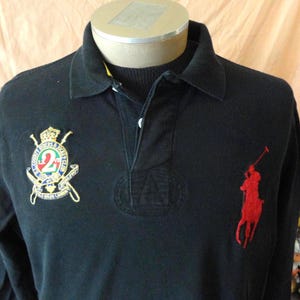 May include: Black long-sleeve polo shirt with a button-down collar. Features a red polo player logo on the chest and an embroidered crest on the upper left chest. The crest includes a crown, the number "2", and the words "County Riders Polo Club".