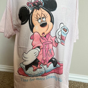 May include: Light pink t-shirt featuring a cartoon Minnie Mouse in a pink robe, curlers, and slippers, holding a cup. The text "Time for more Beauty Sleep" is printed at the bottom in light blue.
