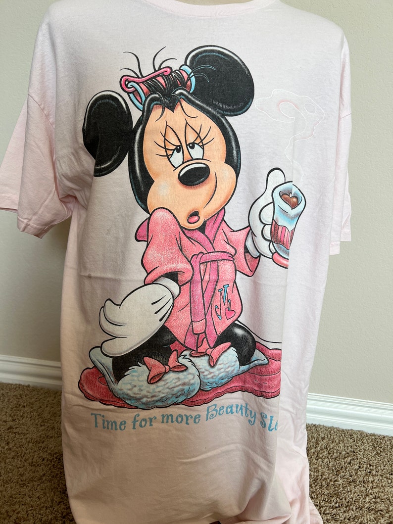 May include: Light pink t-shirt featuring a cartoon Minnie Mouse in a pink robe, curlers, and slippers, holding a cup. The text "Time for more Beauty Sleep" is printed at the bottom in light blue.