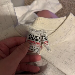 May include: Close-up of a white and gray speckled sweatshirt with a tag that reads "ONEITA POWER-SWL". The tag also indicates the sweatshirt is made of polyester and is size XL (50-52).