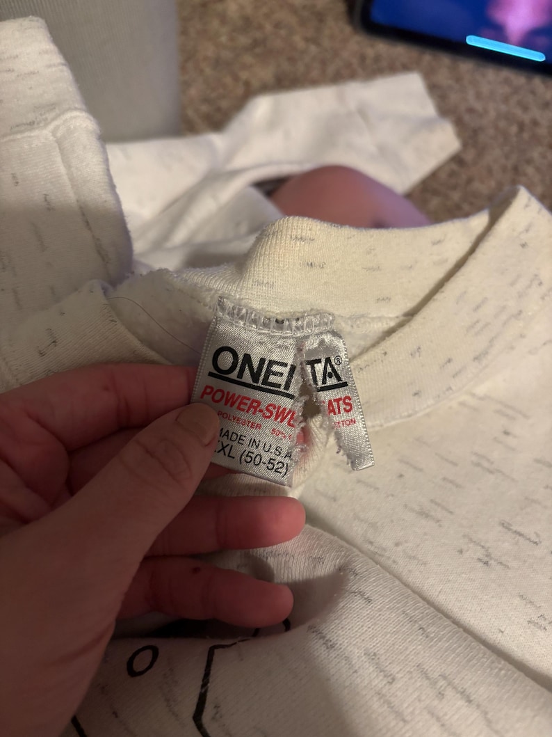 May include: Close-up of a white and gray speckled sweatshirt with a tag that reads "ONEITA POWER-SWL". The tag also indicates the sweatshirt is made of polyester and is size XL (50-52).