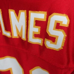以下が含まれることがあります： 赤色のジャージのクローズアップ。名前「HOLMES」は、黄色のトリムが付いた白いブロック文字で表示されています。番号「15」が下部に部分的に表示されています。ジャージはリブ編みのテクスチャです。