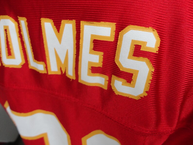 以下が含まれることがあります： 赤色のジャージのクローズアップ。名前「HOLMES」は、黄色のトリムが付いた白いブロック文字で表示されています。番号「15」が下部に部分的に表示されています。ジャージはリブ編みのテクスチャです。