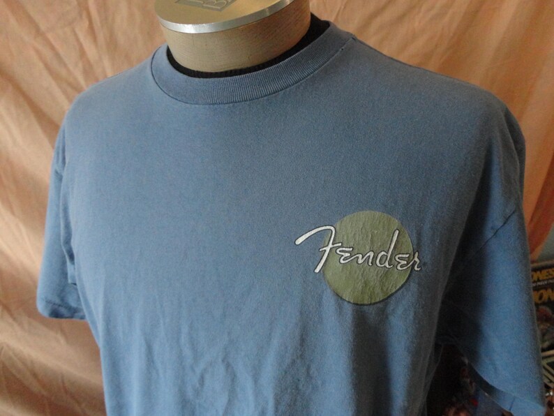 May include: A light blue t-shirt with the Fender logo on the front. The logo is in white script with a green circle behind it. The shirt has short sleeves and a crew neck.