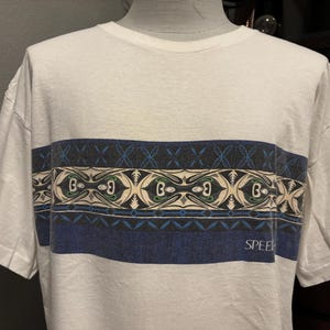 Pode incluir: Camiseta branca com uma faixa horizontal estampada no peito. A faixa apresenta um design em azul, cinza e bege, com a palavra "SPEED" impressa em azul. A camiseta tem mangas curtas e gola redonda.