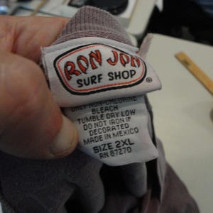 May include: A close-up of a tag from a Ron Jon Surf Shop garment. The tag is white with a red and black logo. The text on the tag includes care instructions and the size 2XL. The garment is a muted purple color.