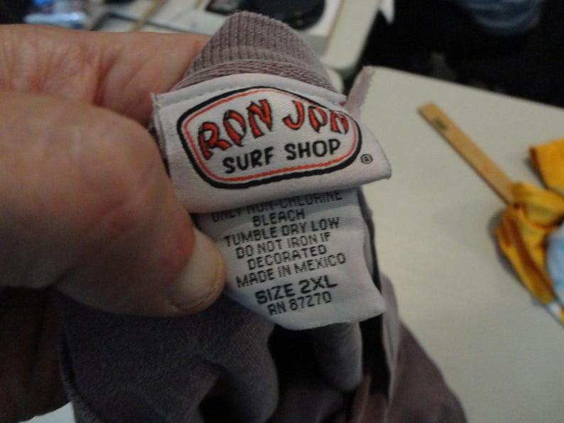 May include: A close-up of a tag from a Ron Jon Surf Shop garment. The tag is white with a red and black logo. The text on the tag includes care instructions and the size 2XL. The garment is a muted purple color.