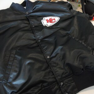 May include: Black satin bomber jacket with a snap front closure. Features a white and red logo patch on the chest. Ribbed cuffs and collar. A classic style.