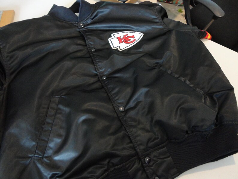 May include: Black satin bomber jacket with a snap front closure. Features a white and red logo patch on the chest. Ribbed cuffs and collar. A classic style.