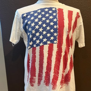 Puede incluir: Camiseta blanca con un gráfico de bandera estadounidense roja, blanca y azul impreso en la parte delantera. La bandera está pintada en un estilo desgastado.