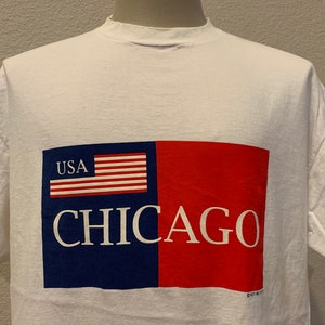 Puede incluir: Camiseta blanca con un gráfico rojo, blanco y azul de la bandera estadounidense y la palabra "CHICAGO" impresa en ella. El gráfico también incluye la palabra "USA" impresa en la bandera.