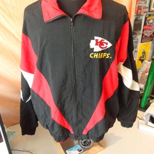 Pode incluir: Casaco preto e vermelho com z&iacute;per e o logotipo dos Kansas City Chiefs no peito. O casaco apresenta gola e detalhes vermelhos, com listras brancas nas mangas. O logotipo dos Chiefs &eacute; branco com uma seta vermelha e a palavra "CHIEFS" em amarelo.