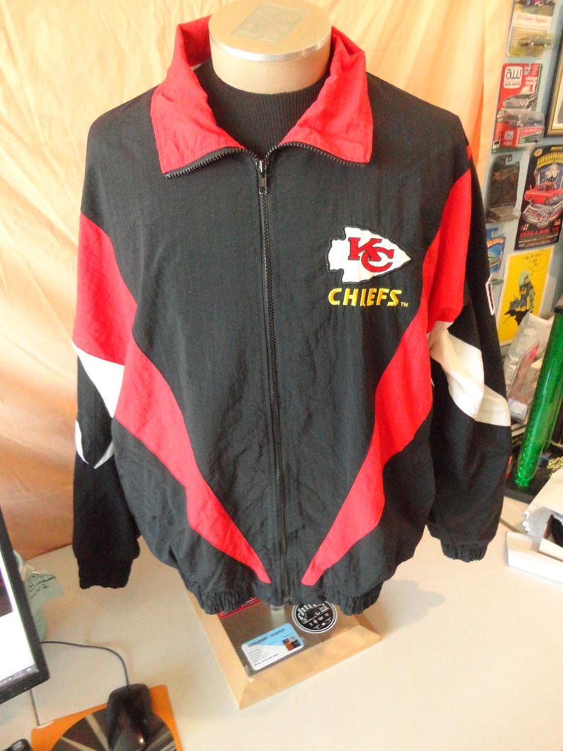Pode incluir: Casaco preto e vermelho com z&iacute;per e o logotipo dos Kansas City Chiefs no peito. O casaco apresenta gola e detalhes vermelhos, com listras brancas nas mangas. O logotipo dos Chiefs &eacute; branco com uma seta vermelha e a palavra "CHIEFS" em amarelo.