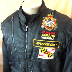 May include: A black jacket featuring a front zipper closure and multiple embroidered patches on the chest. The patches include the Ordnance Corps U.S.A. emblem, three stacked Yamaha logos, a Dunlop logo, and a Maryland state flag patch with text.
